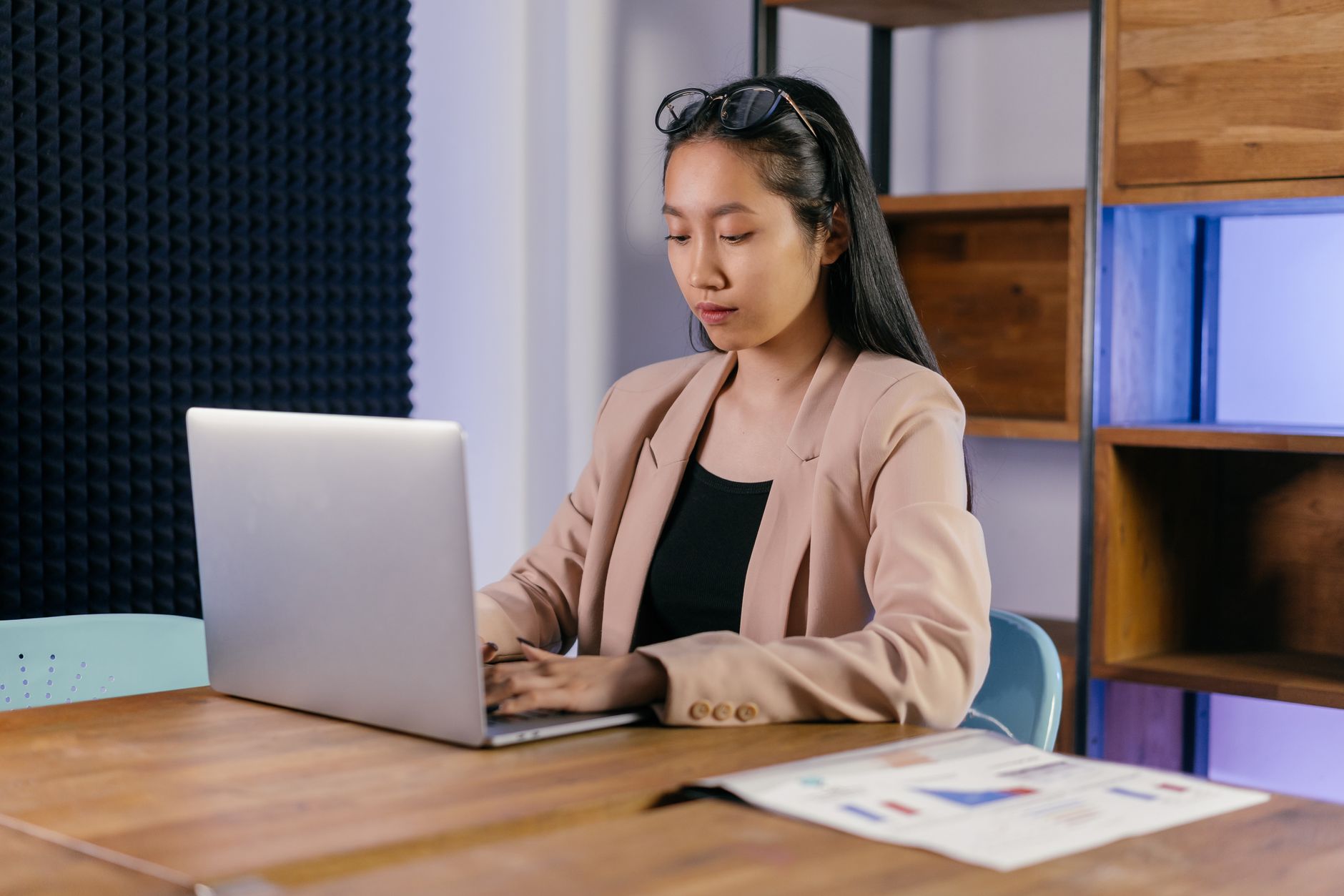 Asian businesswoman in a blazer typing on a laptop in a modern office setting.