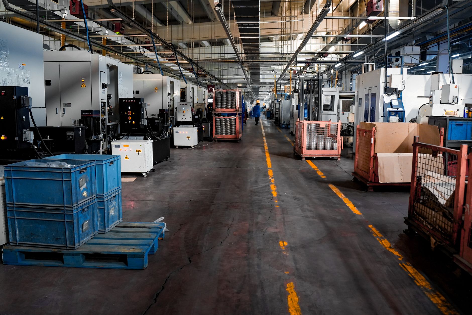 A long, empty aisle inside a manufacturing plant lined with industrial machines, a single worker walking away down the center line