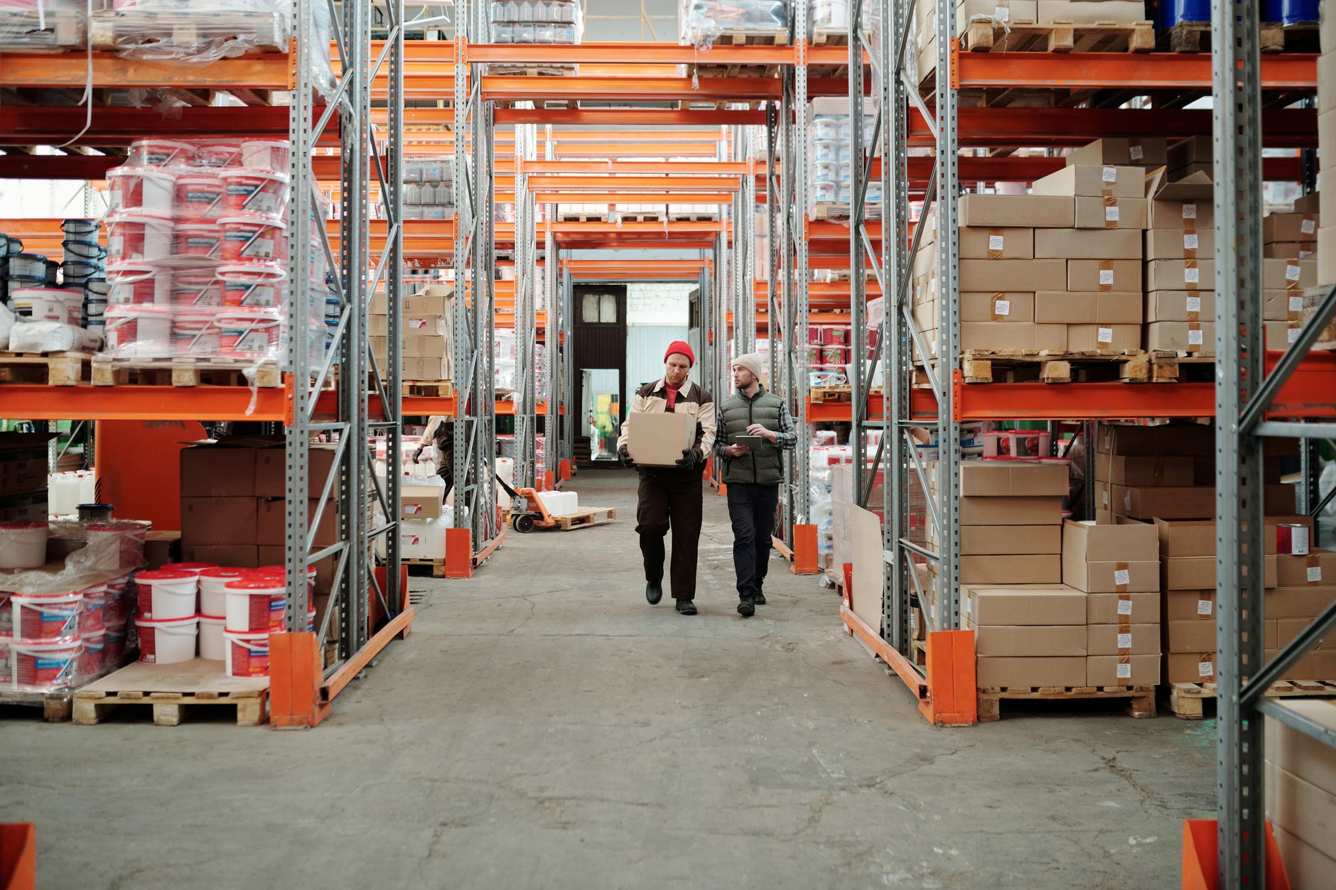 Two warehouse workers walking together down an aisle of tall orange racking stacked with boxes, one carrying a box and the other checking a tablet