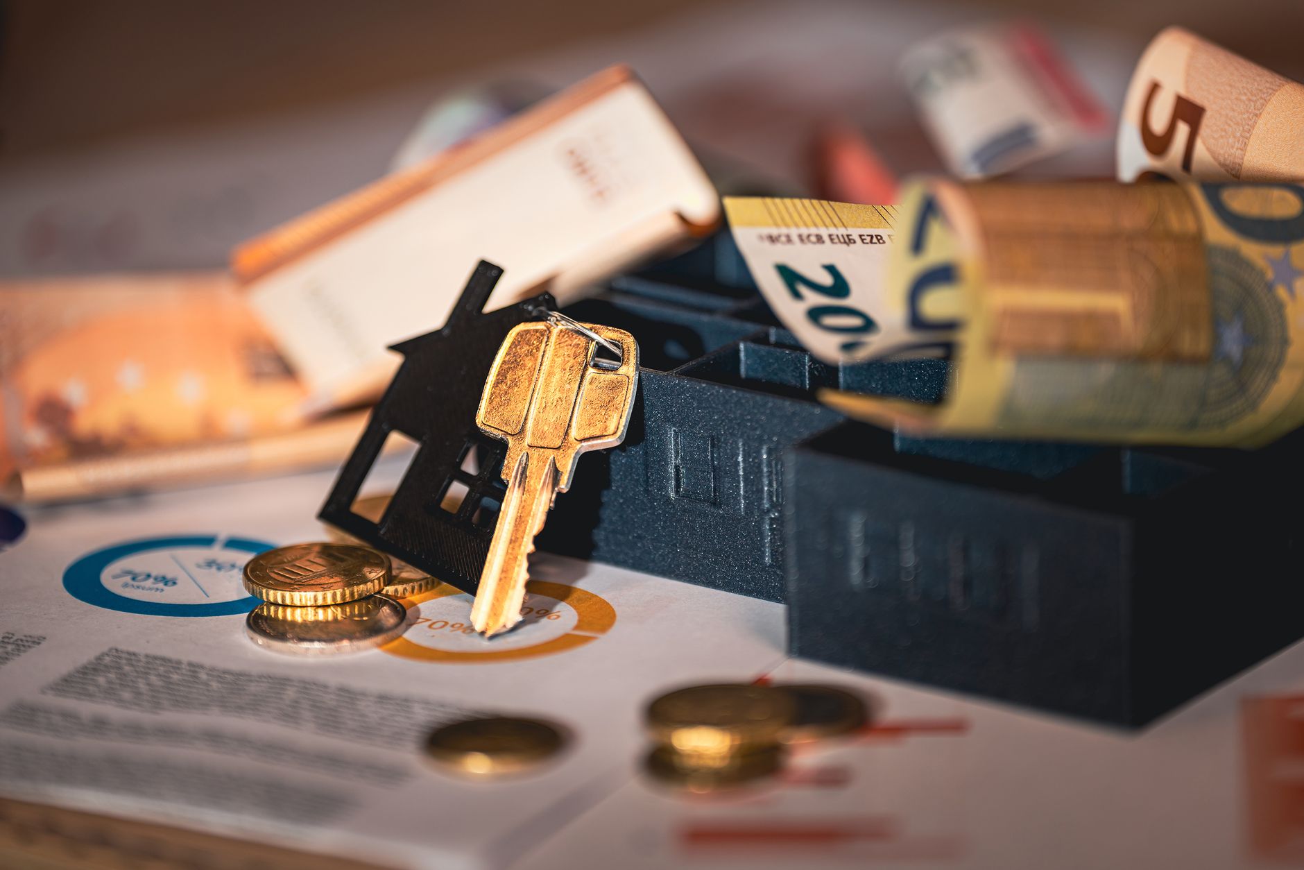 A small model house sitting on a stack of coins and folded paper bills on a wooden desk