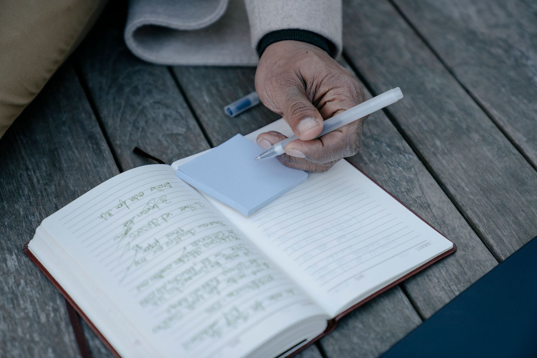 Person writing in a journal, focusing on hand holding pen with notepad on a wooden table.