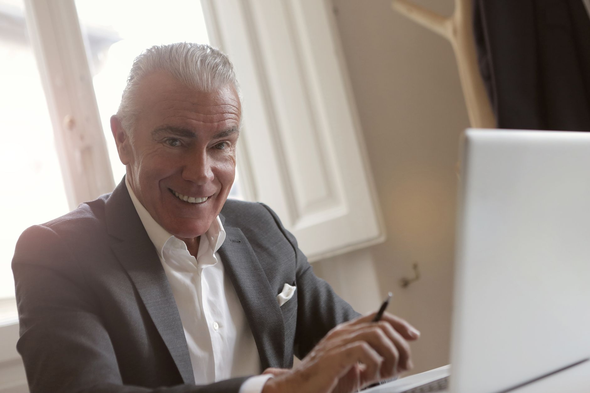 Smiling senior businessman in office using a laptop with a pen in hand, bright light from windows.