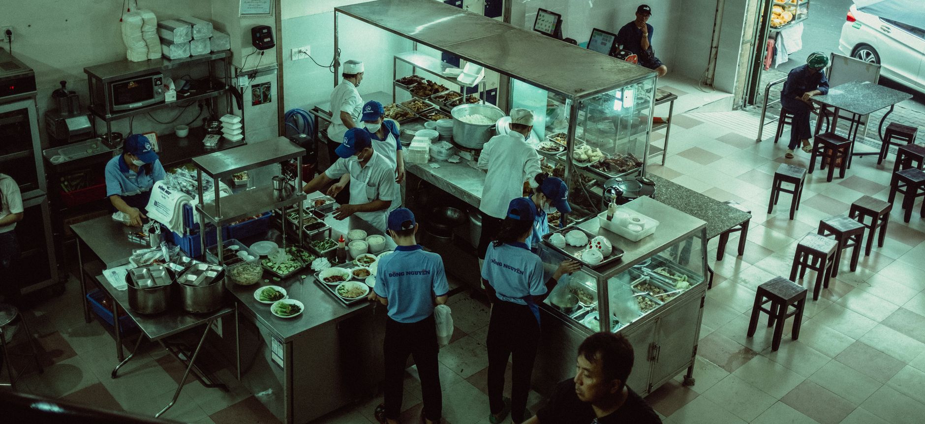 Line cook in a black apron assembling a bowl on a stainless steel prep counter, hands moving between metal containers of rice, beans and salsa
