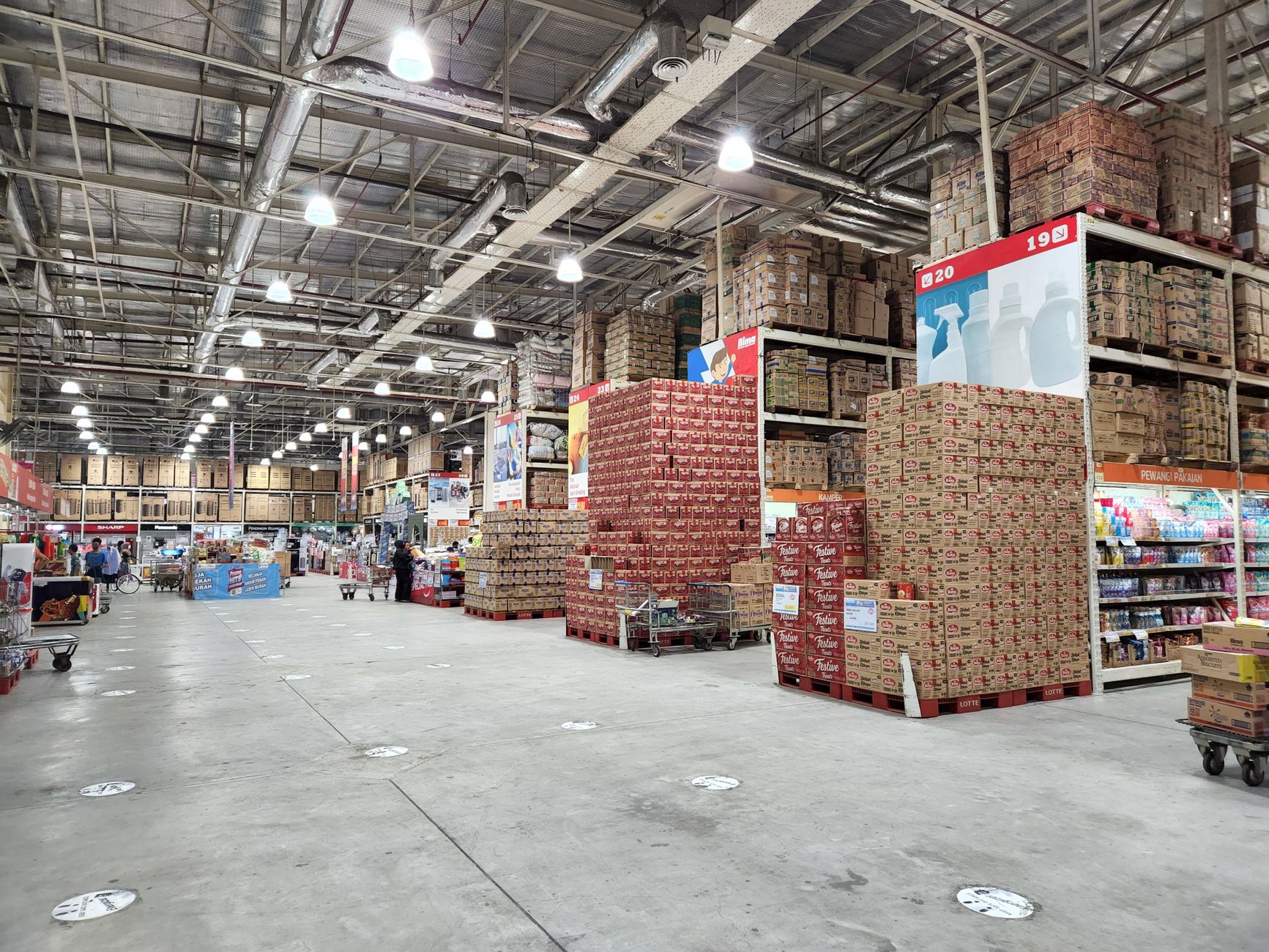 Wide aisle of a warehouse-style store with tall steel racking, pallets of boxed goods, and shoppers pushing oversized carts