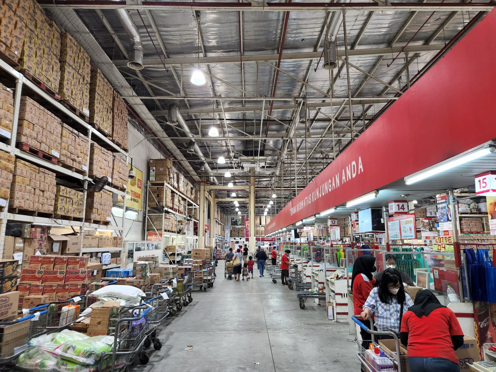 Two employees in a stockroom talking beside a pallet jack and shrink-wrapped inventory, one holding a clipboard