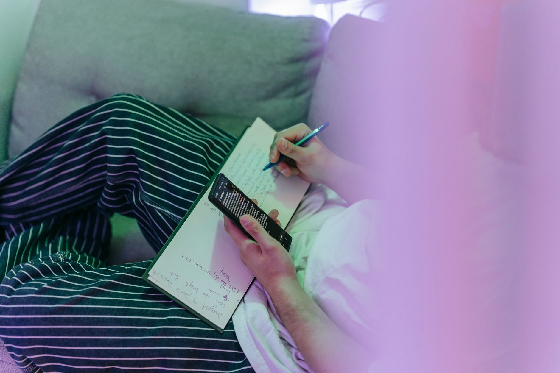 A cozy scene of a person writing notes in a notebook while using a smartphone.