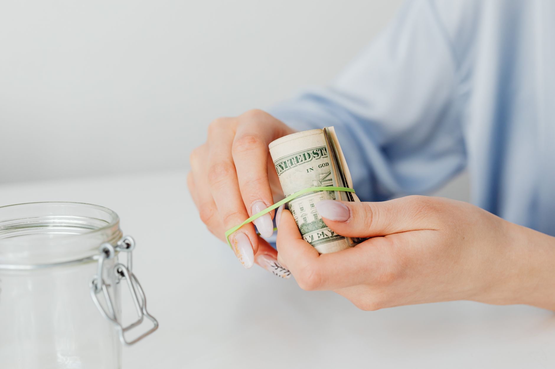 Hands securing rolled US dollar bills beside a savings jar