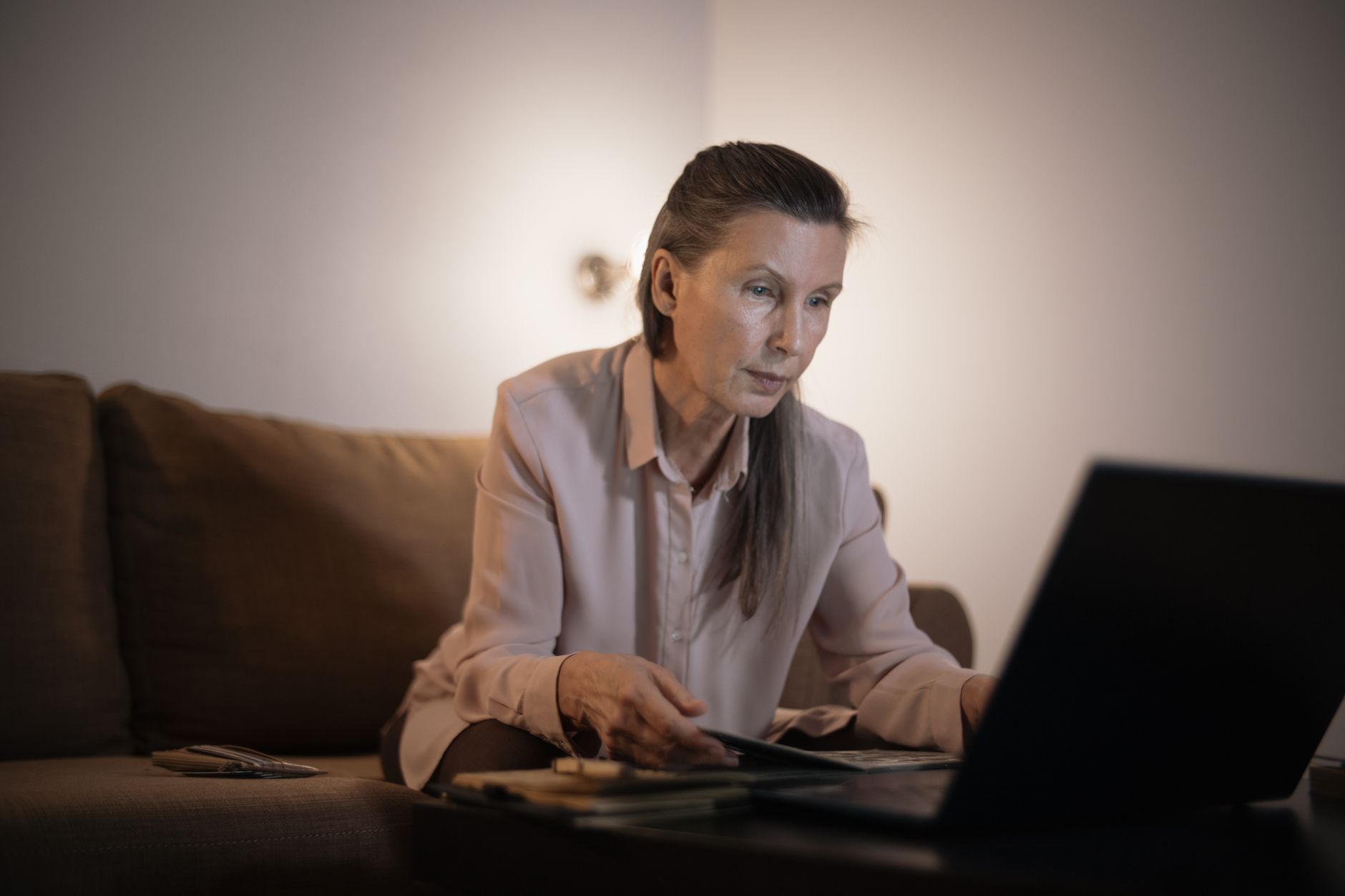 一位女性深夜坐在沙發上,一邊翻看筆記一邊對著筆電工作,神情專注而疲憊