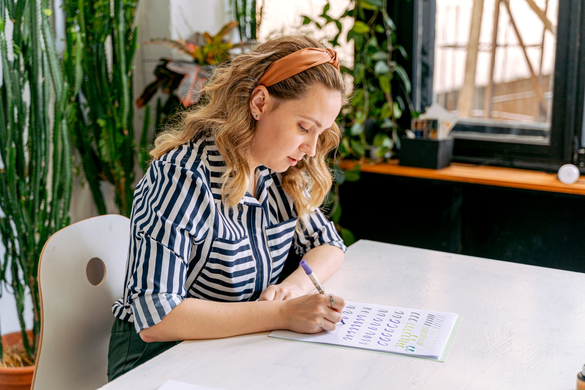 A person writing notes at a desk beside an open notebook and laptop, mid-thought