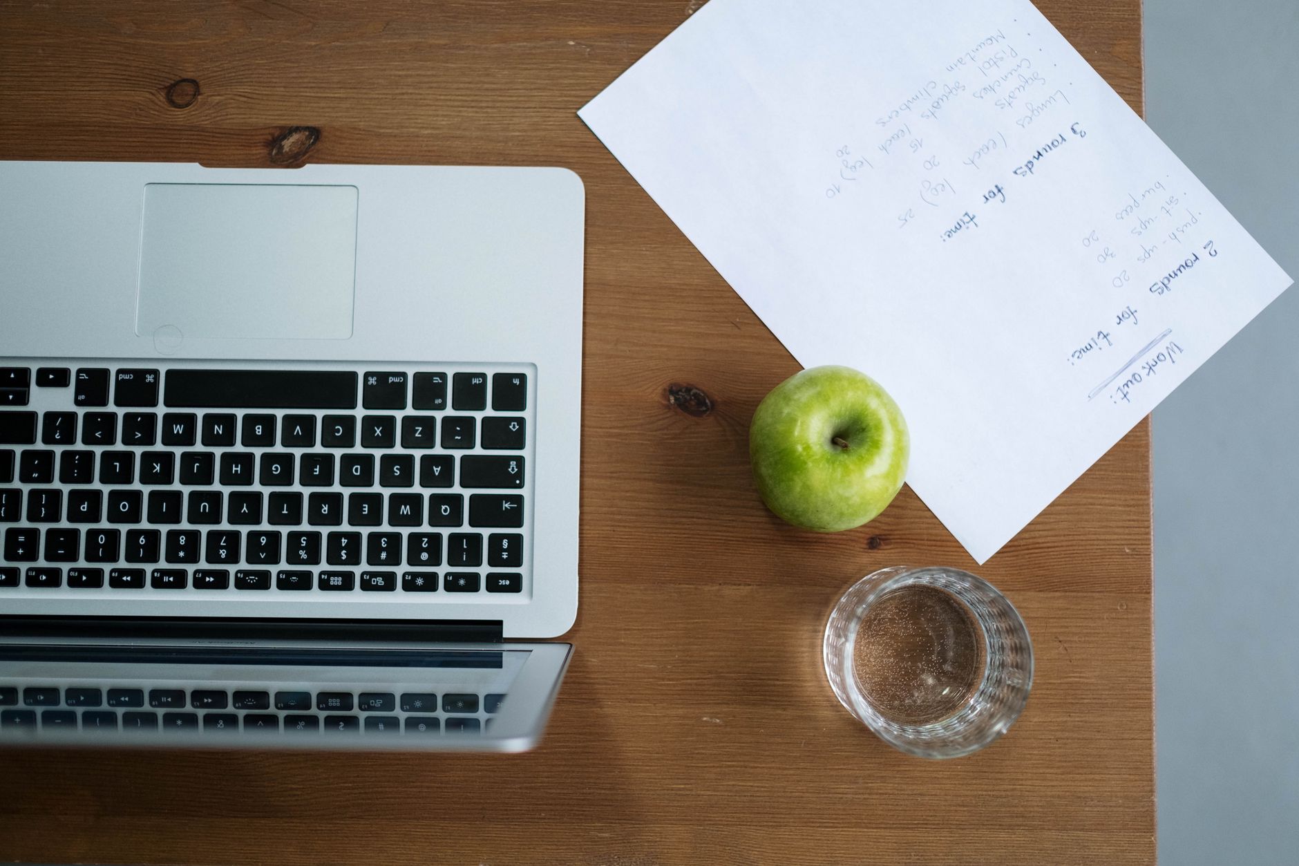 Top view of a workspace with a laptop, green apple, water glass, and paper notes.