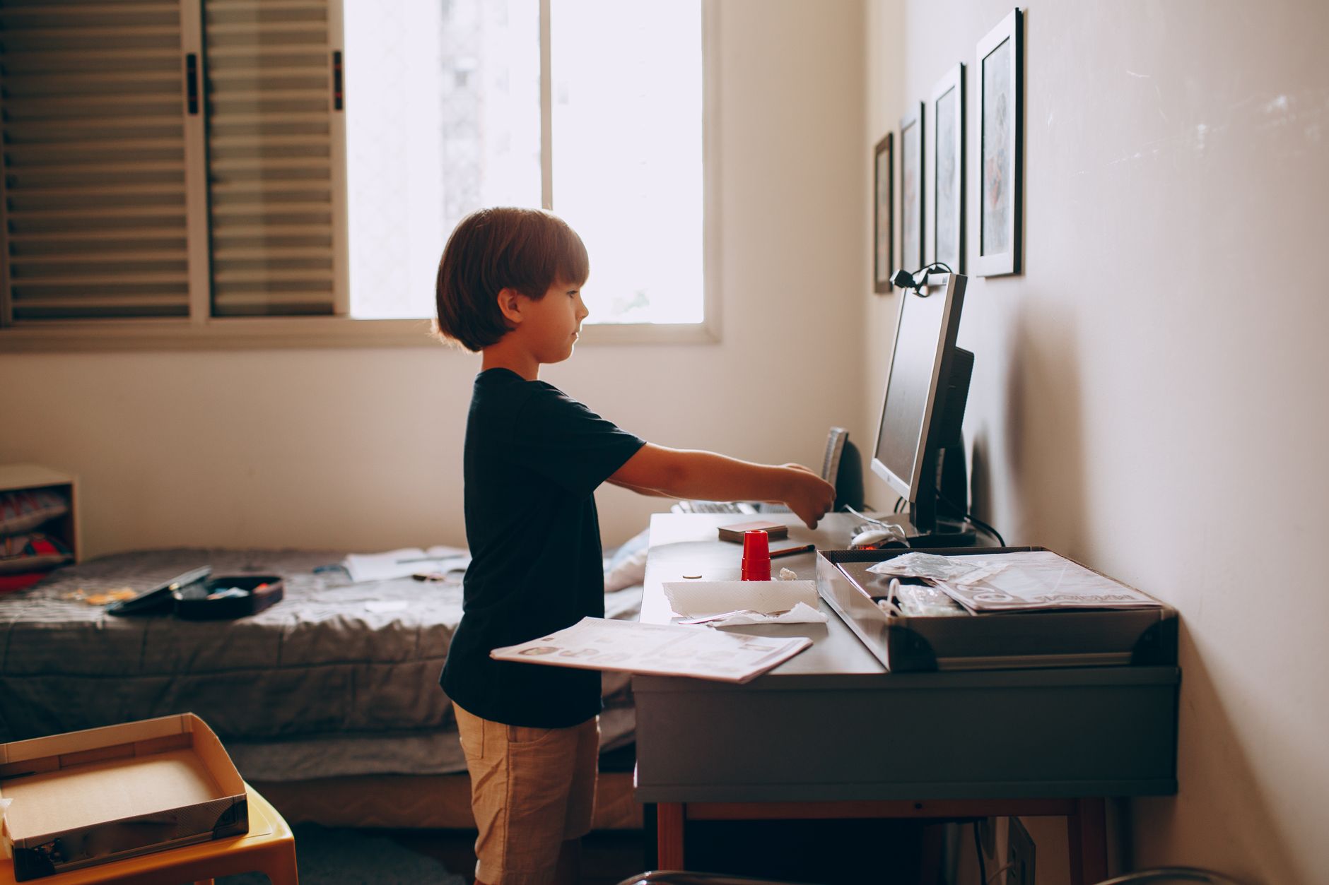 Young boy engaging in activities at a desk in well-lit room with framed pictures.