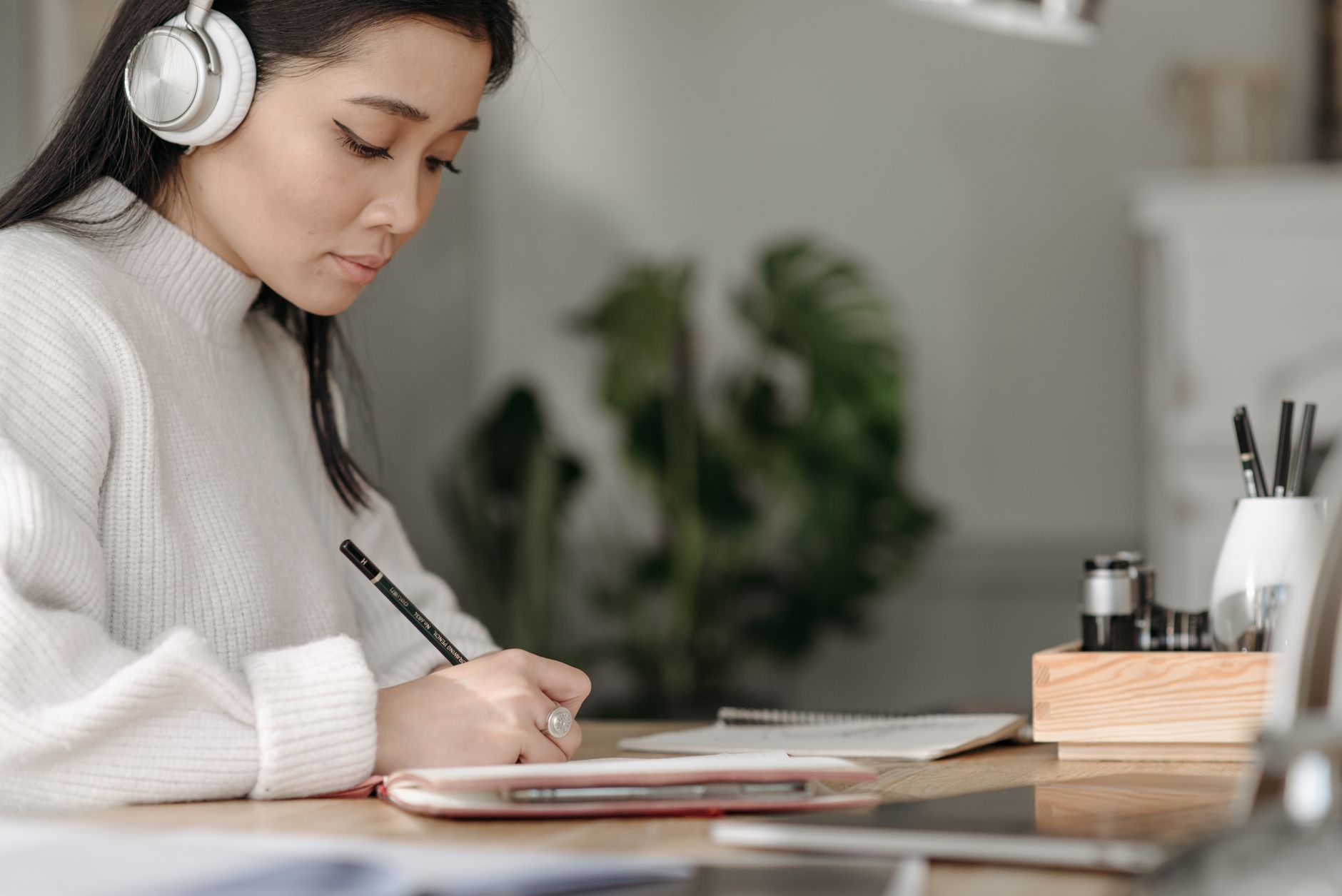 A woman wearing white over-ear headphones writes by hand in a notebook, her phone tucked face-down into the notebook's cover instead of in her hand