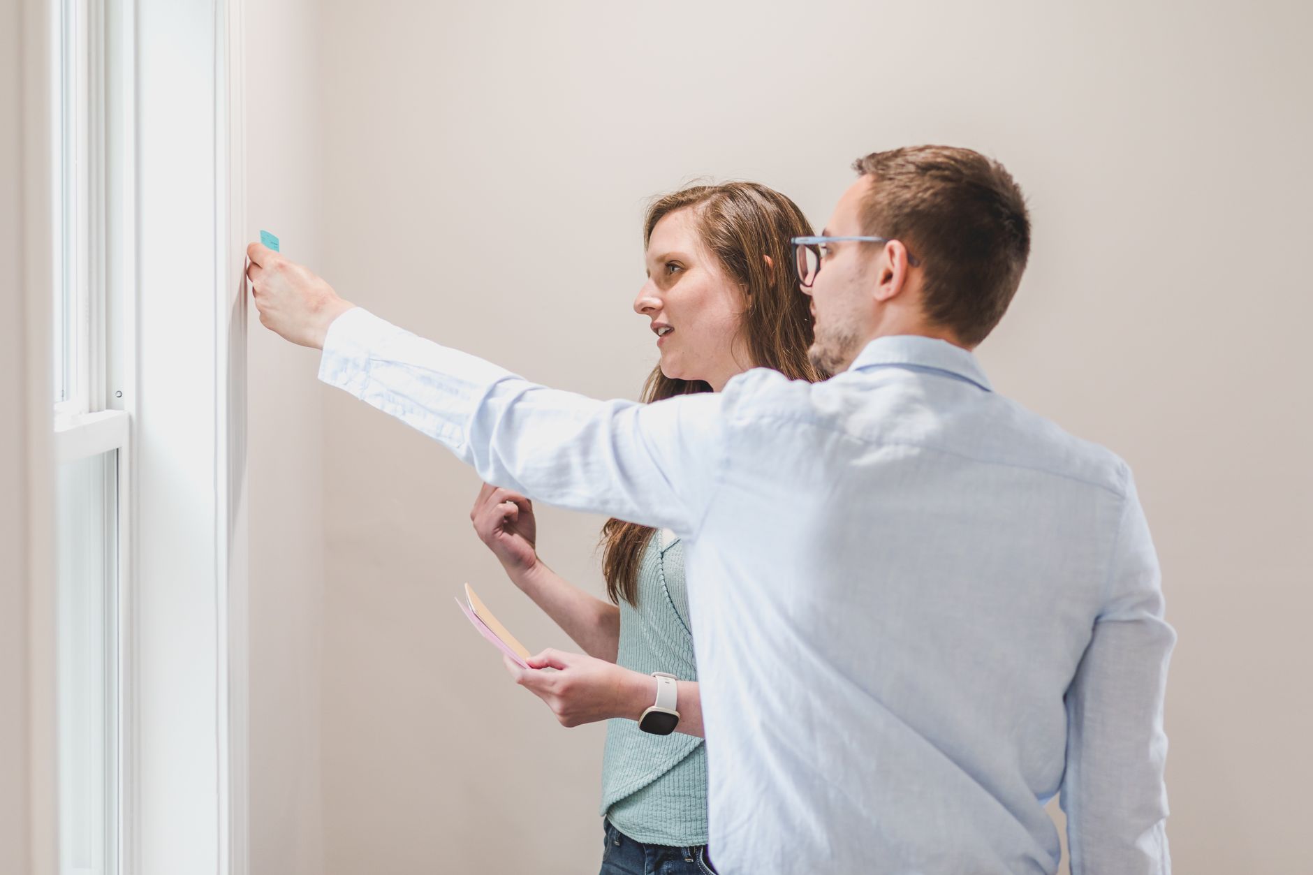 Two coworkers by a window, one carefully placing a single sticky note high on the wall while the other watches