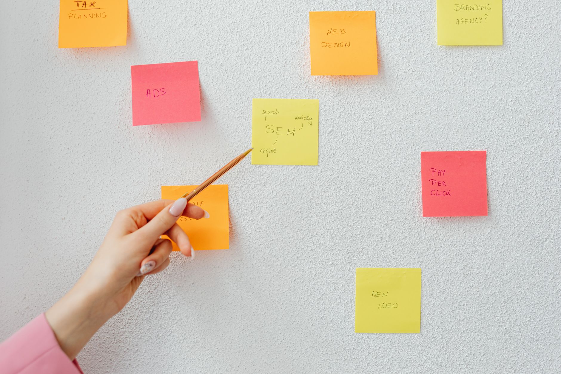 A hand holding a pen beside a wall covered in colorful sticky notes labeled with unrelated tasks like ads, web design, and tax planning