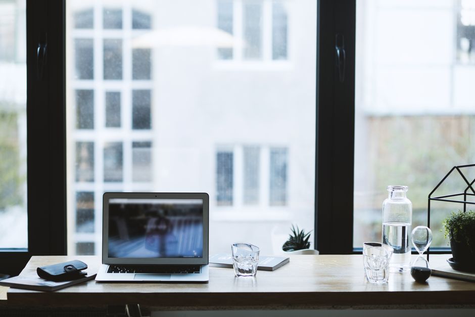 A laptop and glass of water sit on a clear desk near a window.