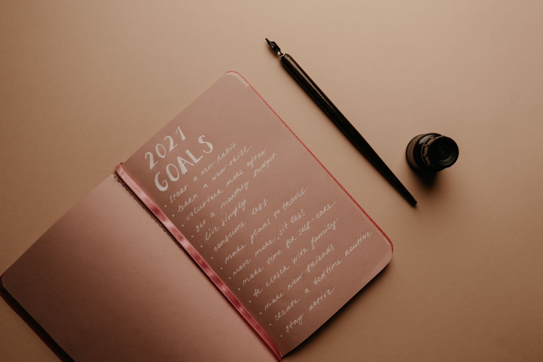 A dip pen resting beside a bottle of ink and a handwritten goals list in an open notebook, photographed at an angle