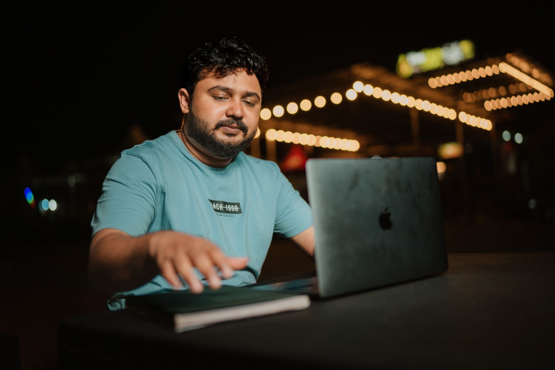 A man working late outdoors on a MacBook, a closed notebook beside him on the table, string lights glowing in the dark background