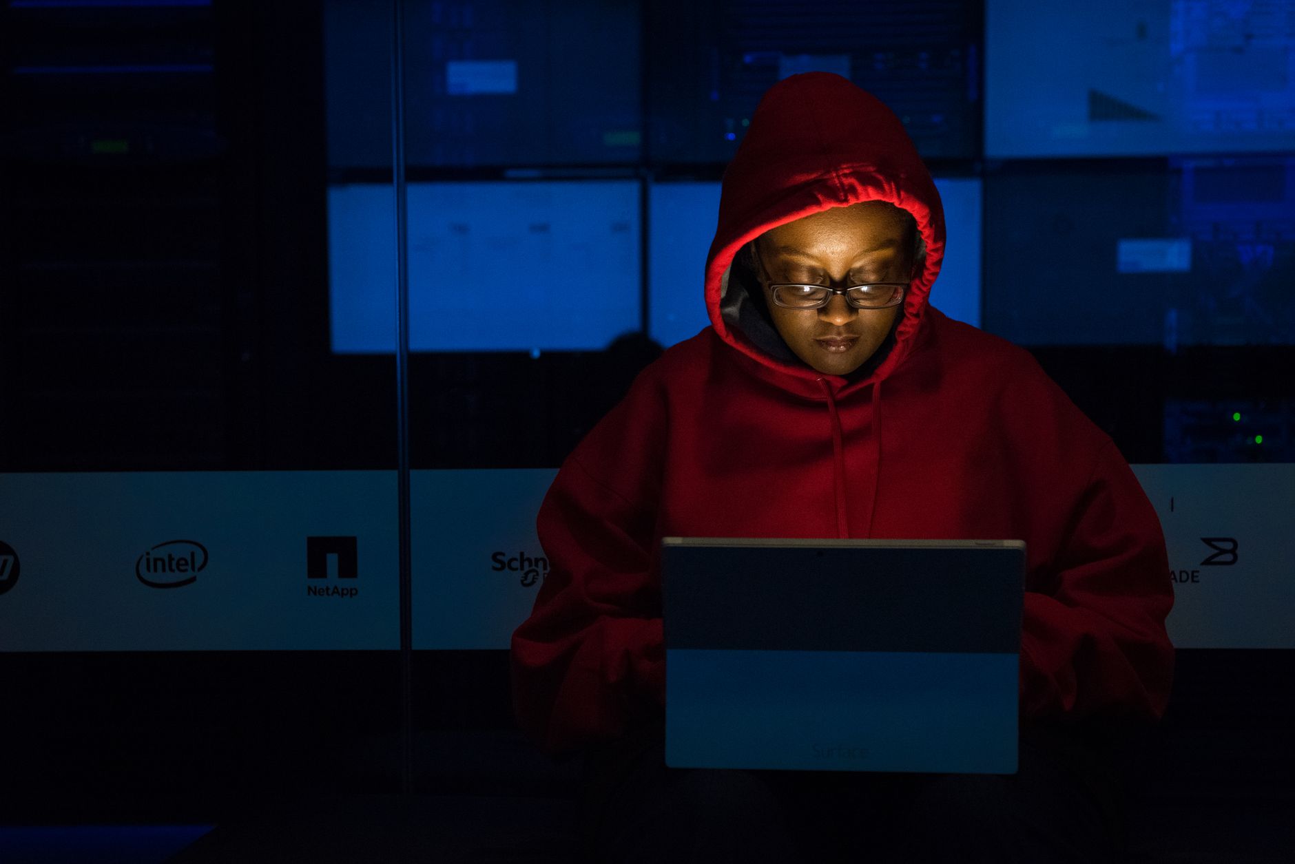 A person in a red hoodie lit only by their laptop screen, working alone at night in front of a wall of server racks and status lights