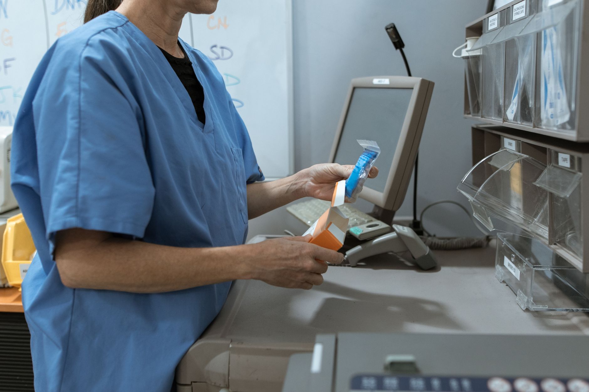 Two healthcare workers in scrubs talking quietly at a clinic reception desk while a patient waits in the background