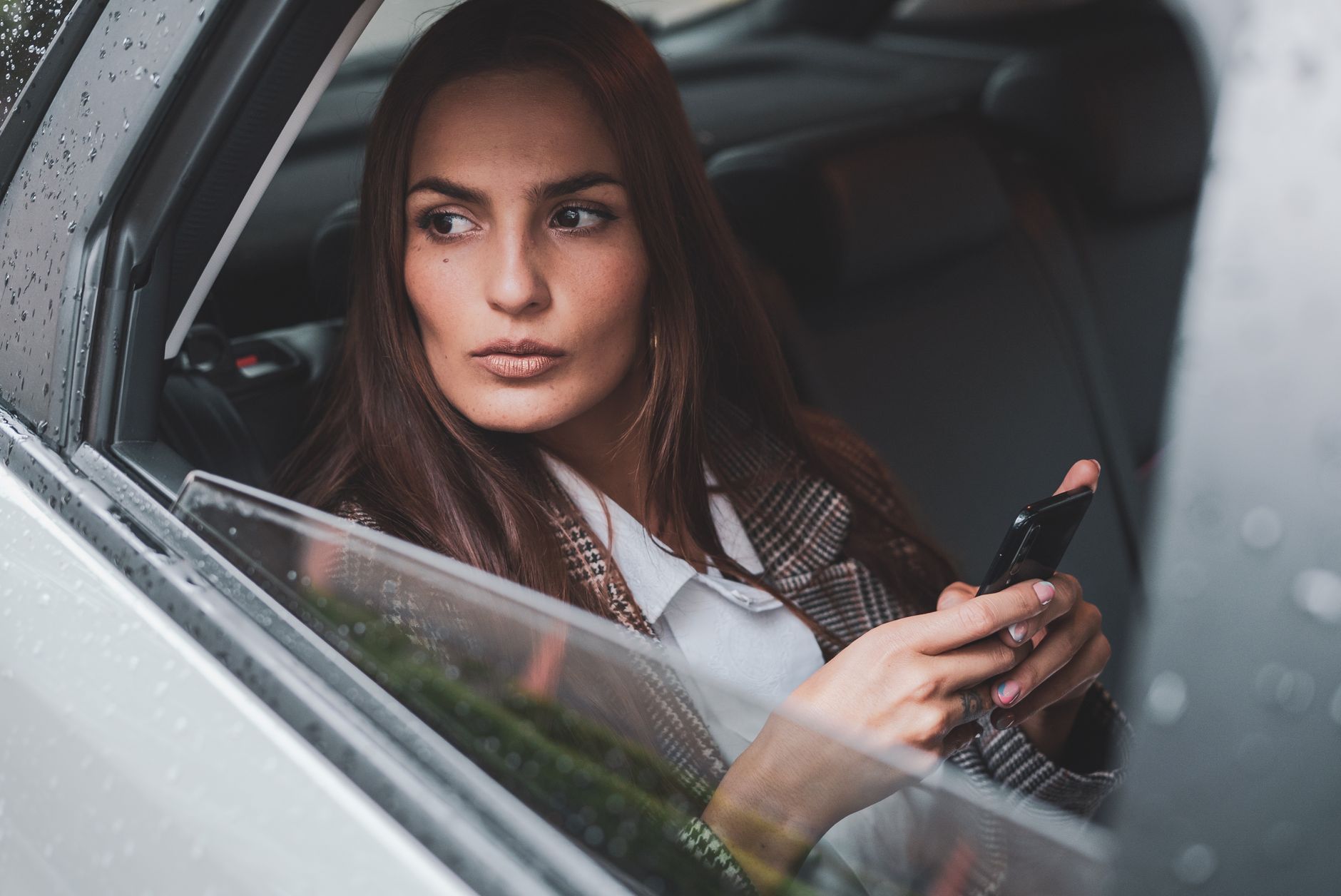 A thoughtful woman looking out of a car window while holding a smartphone, portraying a modern lifestyle.