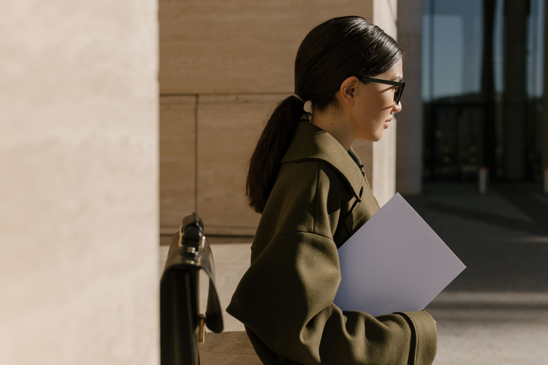 Side view of a woman in an olive coat holding a document, standing outdoors.