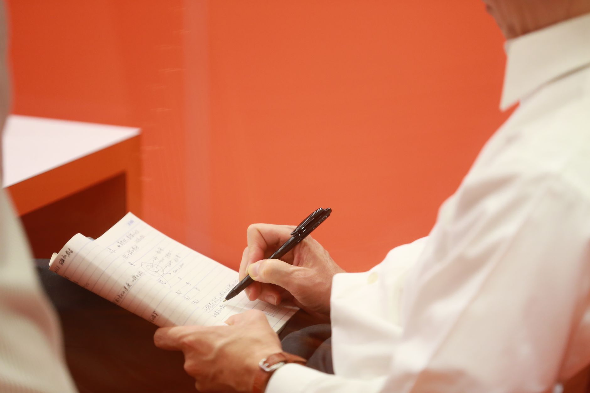 Adult engaged in writing notes with a pen in a notebook against an orange background.