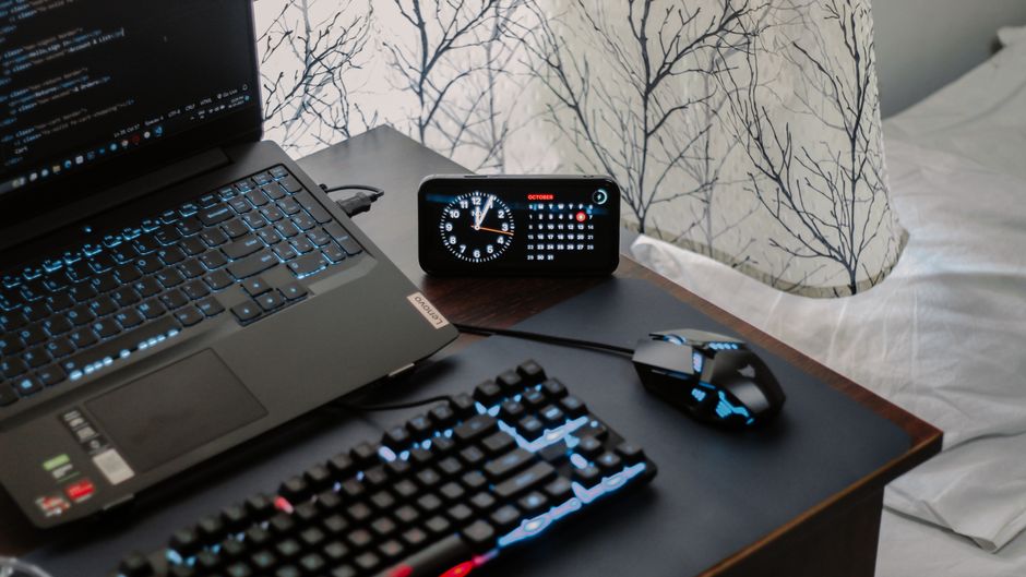 A laptop, keyboard, mouse, and digital clock on a work desk.