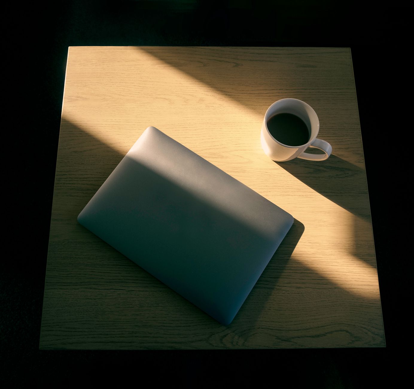 A closed laptop and a white mug of coffee on a wooden table, lit by a single sharp beam of morning sun