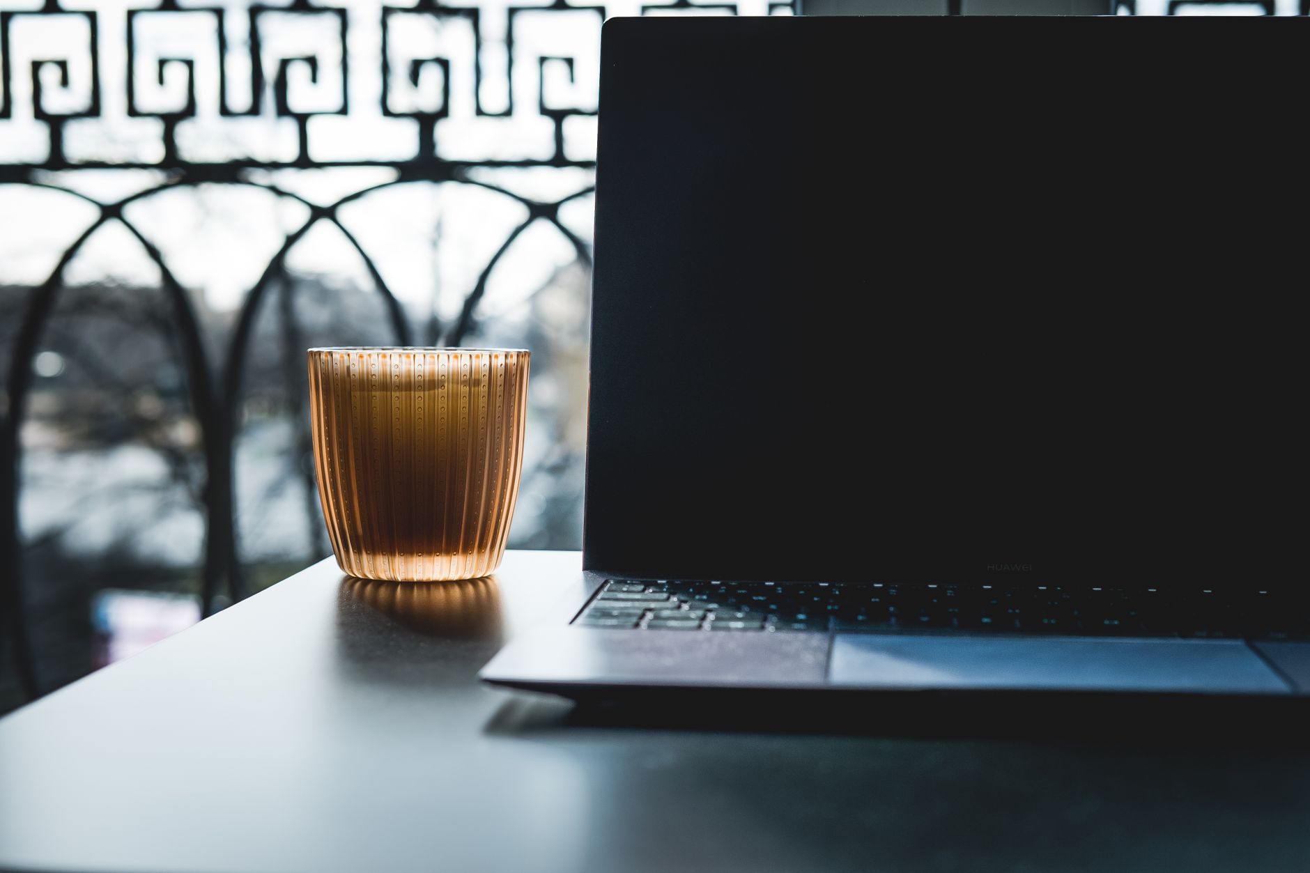 A laptop with its screen dark sits closed beside a glowing cup on a table in front of an ornate iron window at dusk