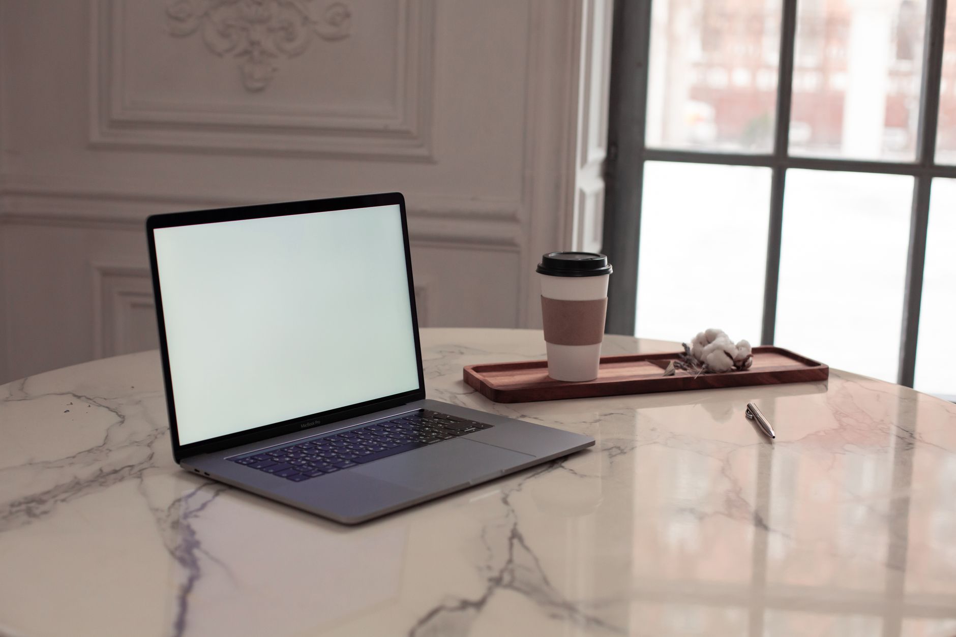 An open laptop with a blank, bright screen on a marble table beside a takeaway coffee cup and a sprig of cotton