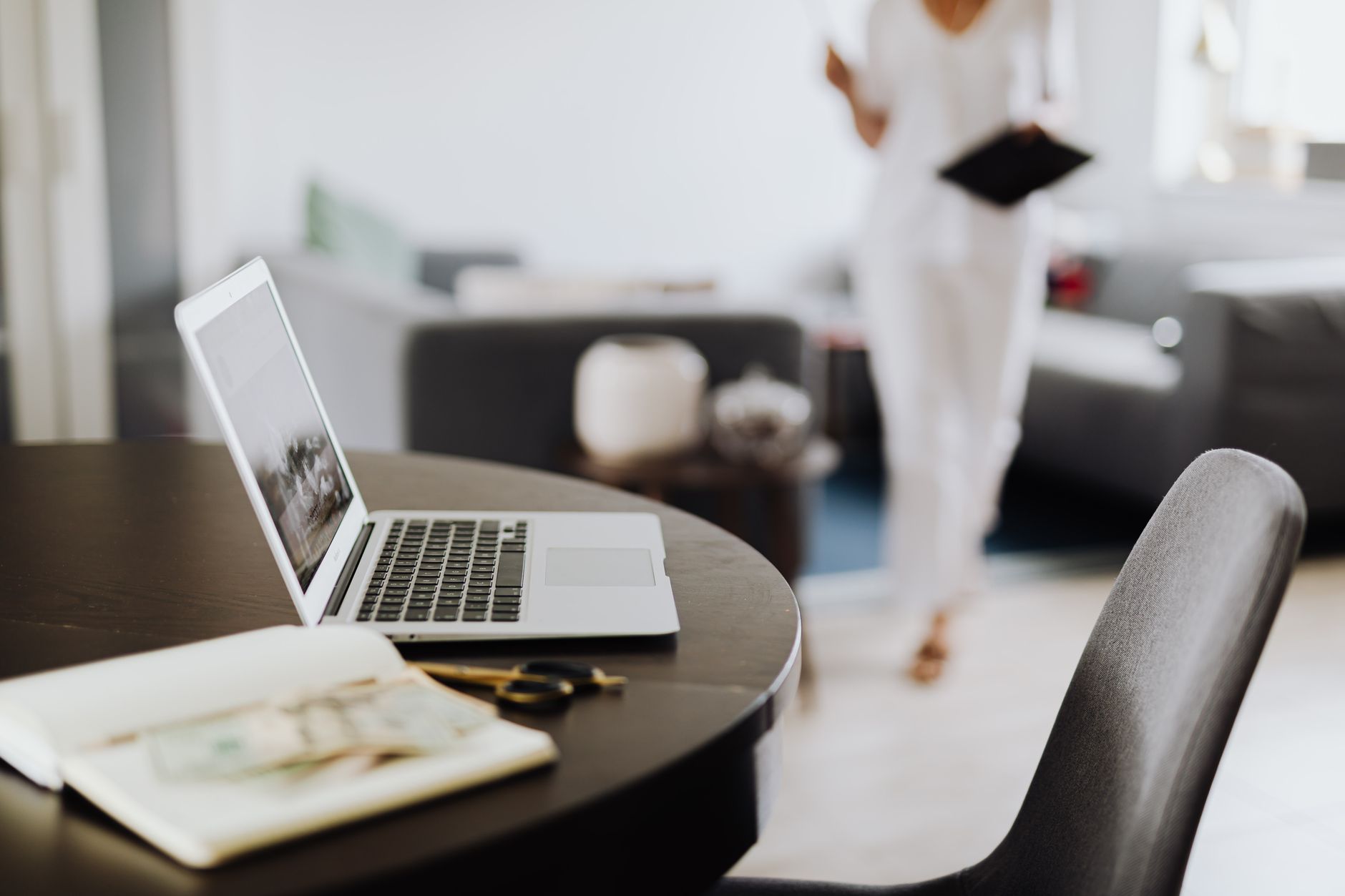An open laptop and notebook on a table in the foreground, a blurred figure walking past with a tablet in the background