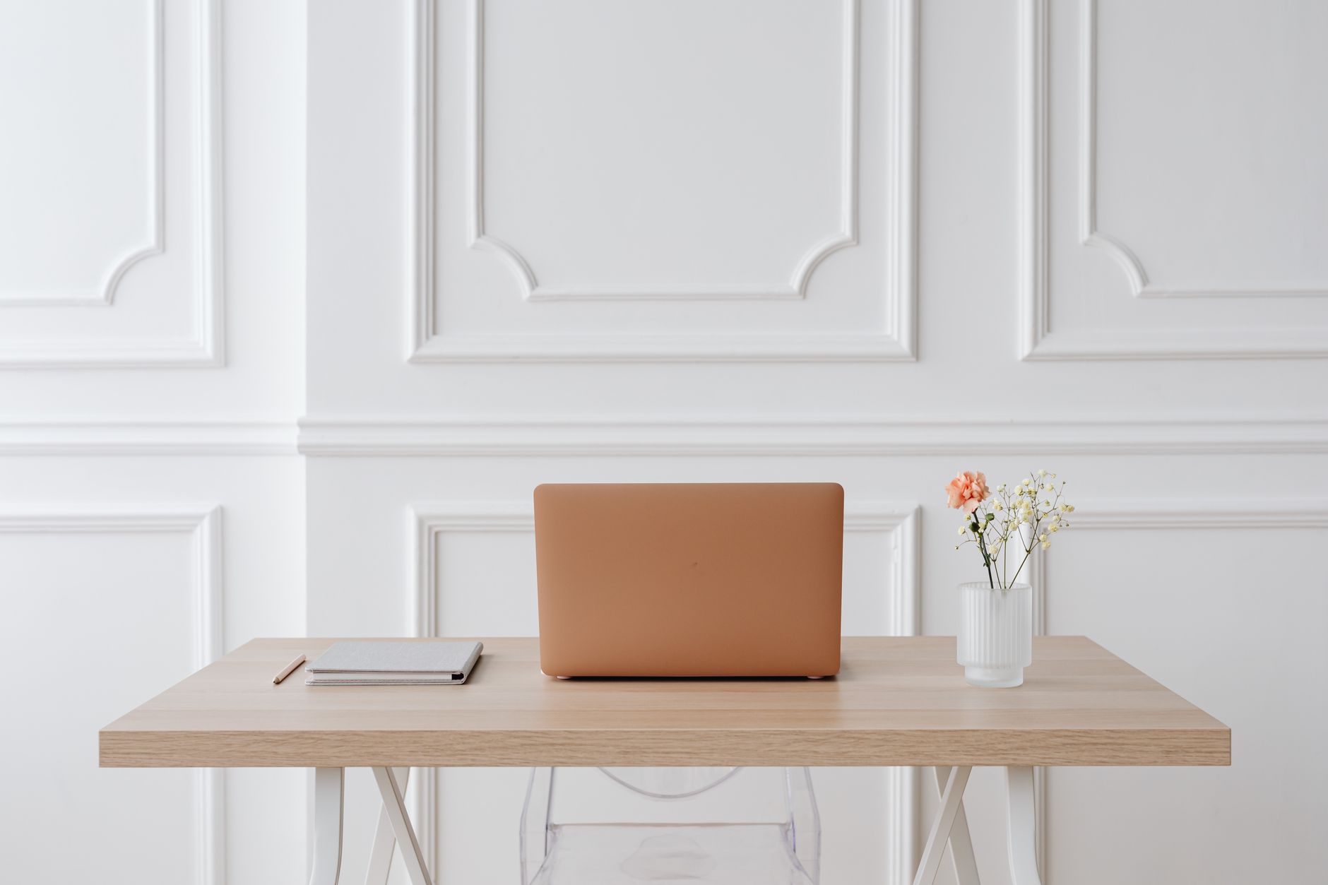 A closed laptop, a small notebook, a pen, and a vase of flowers on an otherwise empty desk