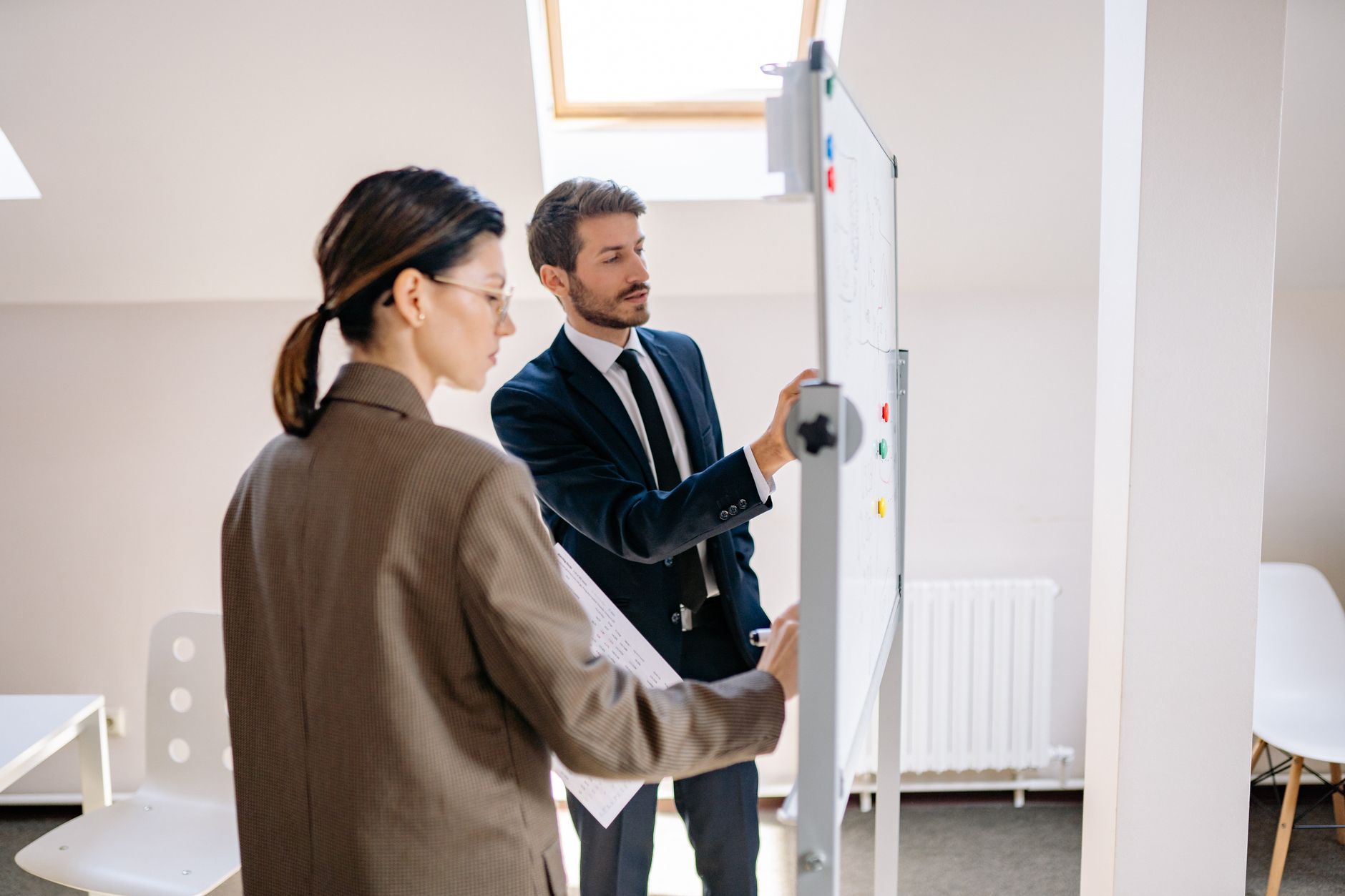 A project manager standing at a whiteboard covered in sticky notes, mid-sentence, explaining a plan to two seated colleagues