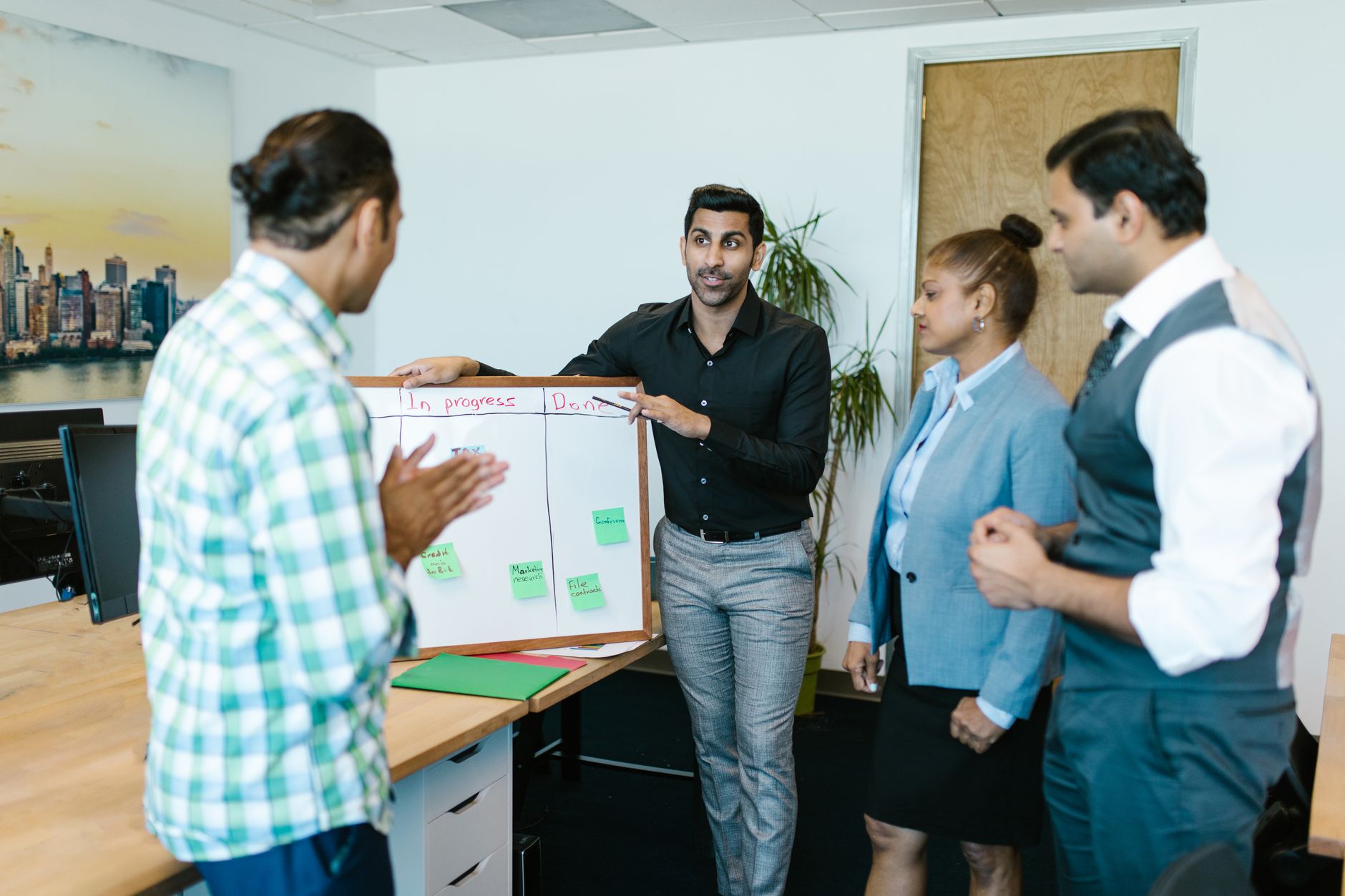 Two people in a small meeting room reviewing a printed timeline on a table, one pointing at a specific milestone