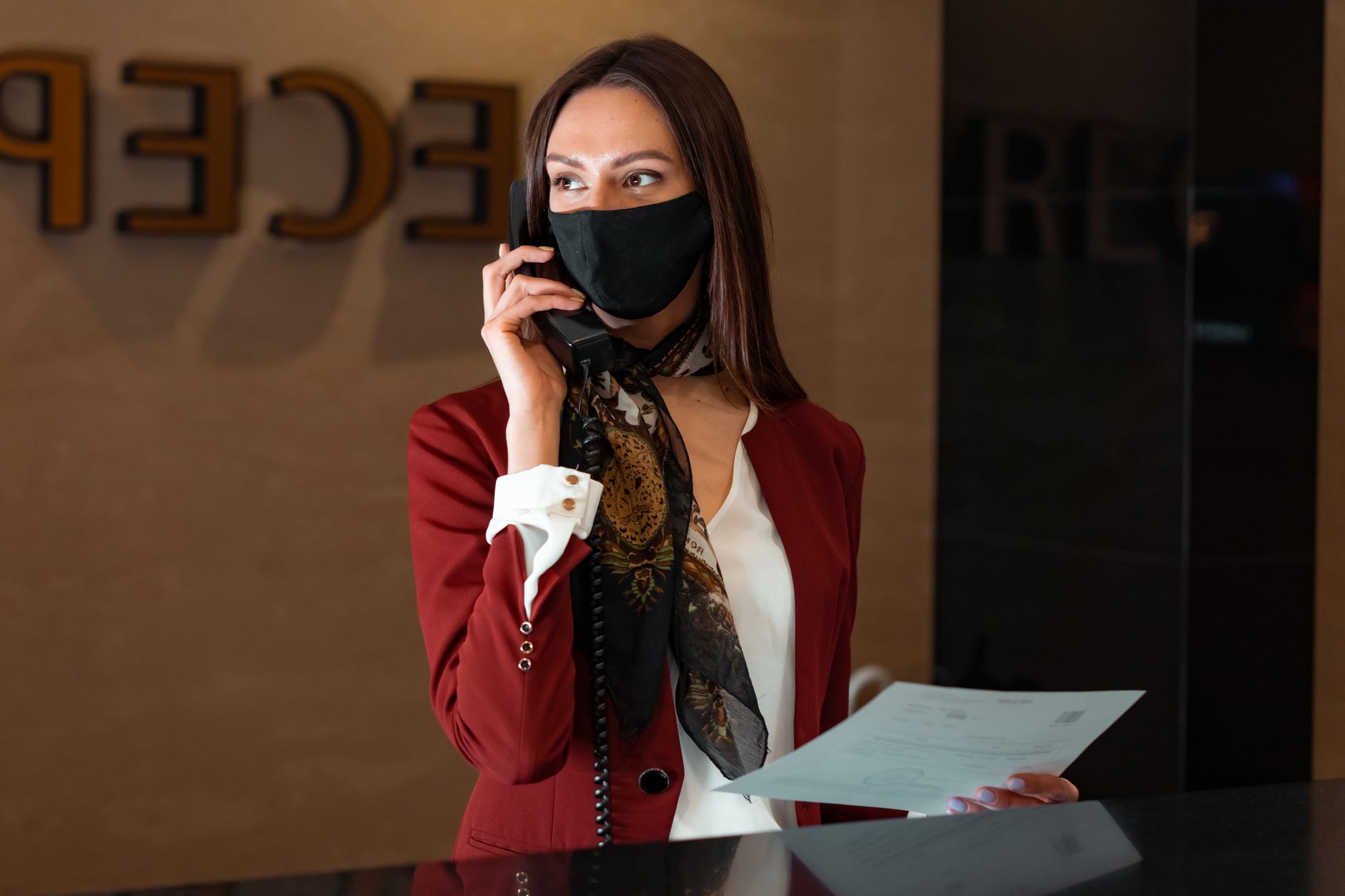 A receptionist at a front desk speaking on a headset while a visitor waits nearby in a bright office lobby