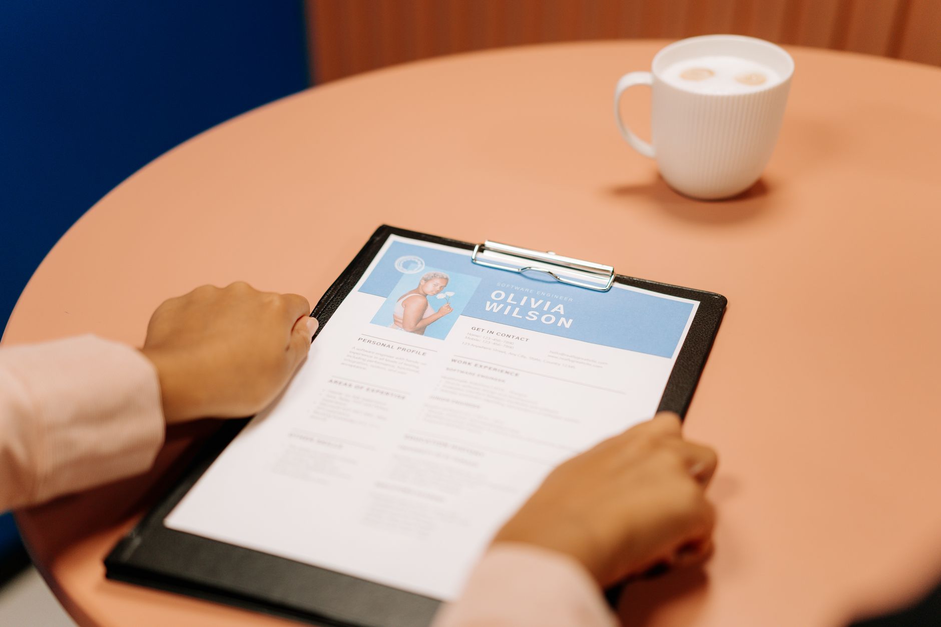 A printed resume on a clipboard showing a photo, name, and personal profile section at the top, next to a cup of coffee