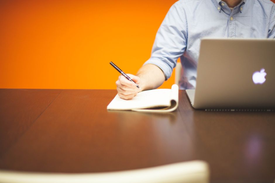 A worker taking notes beside an open laptop on a desk.