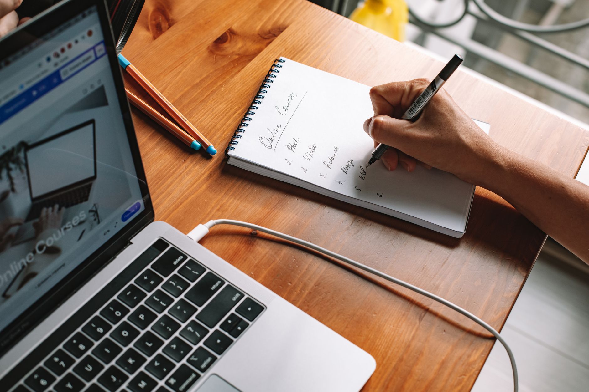 A person at a plain wooden desk writing a short list in a notebook beside a closed laptop