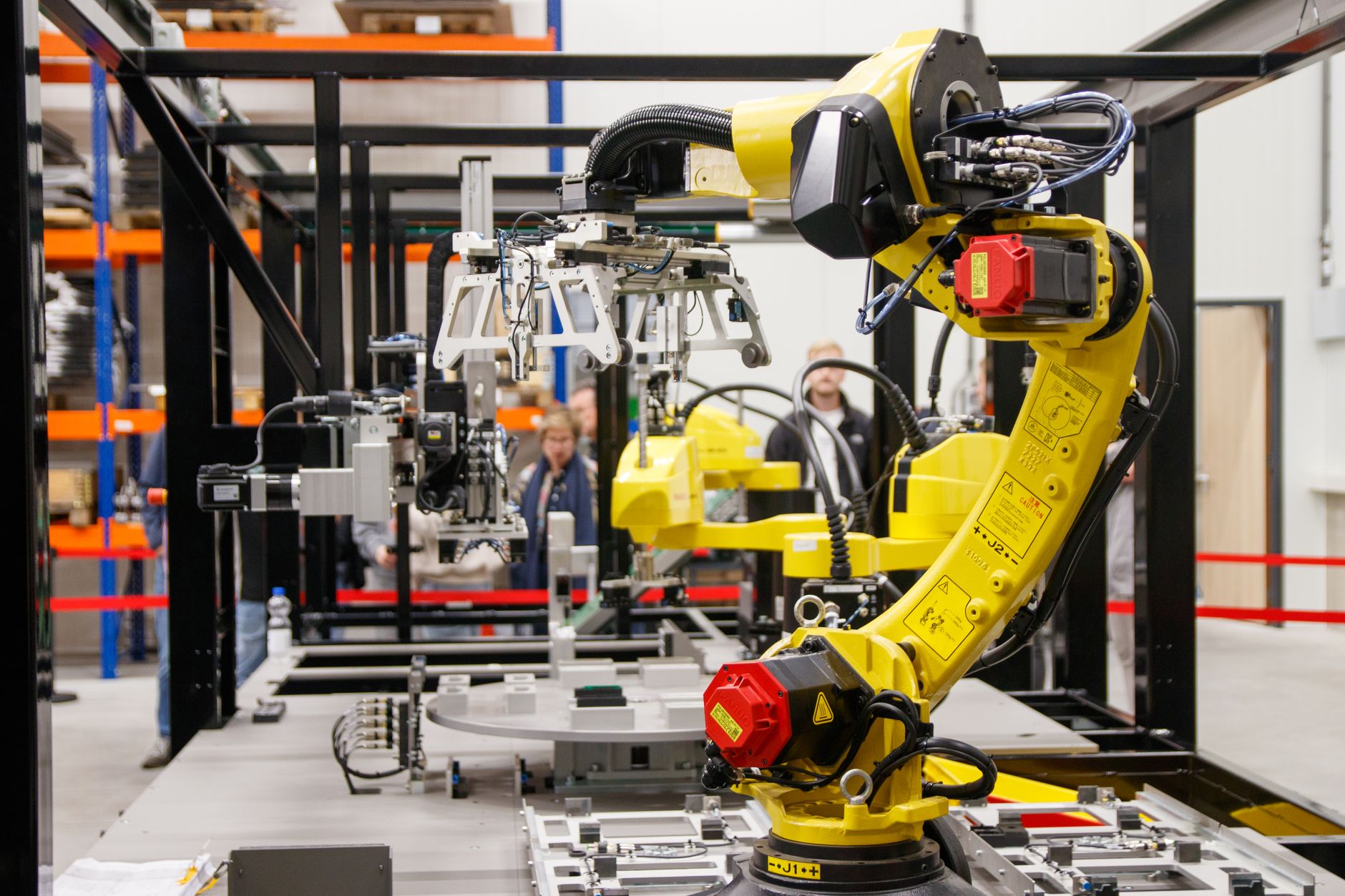 A yellow industrial robotic arm reaching over an assembly line inside a factory, with people observing from the aisle behind it
