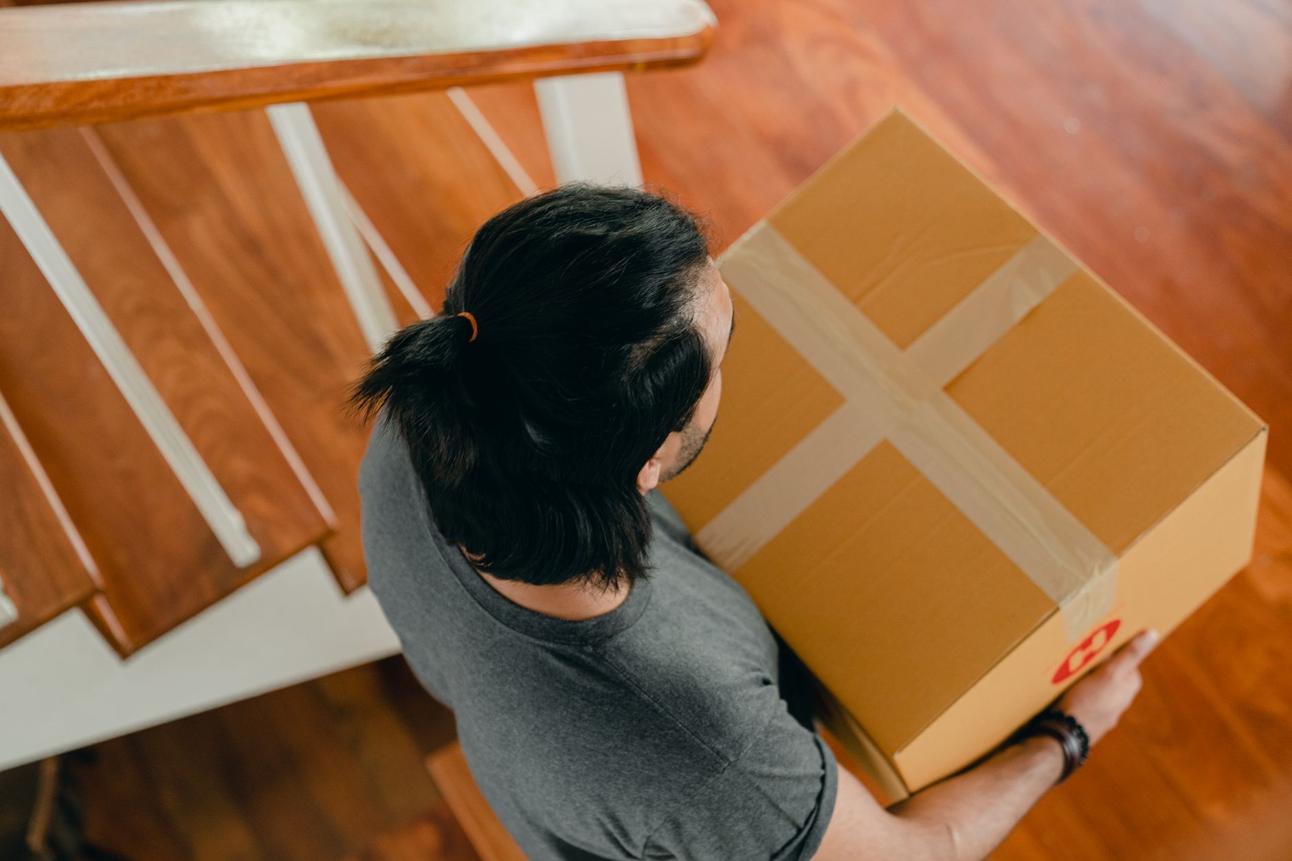 From above of young man with dark hair in casual clothes standing near wooden stairs and carrying carton box during relocation into new apartment