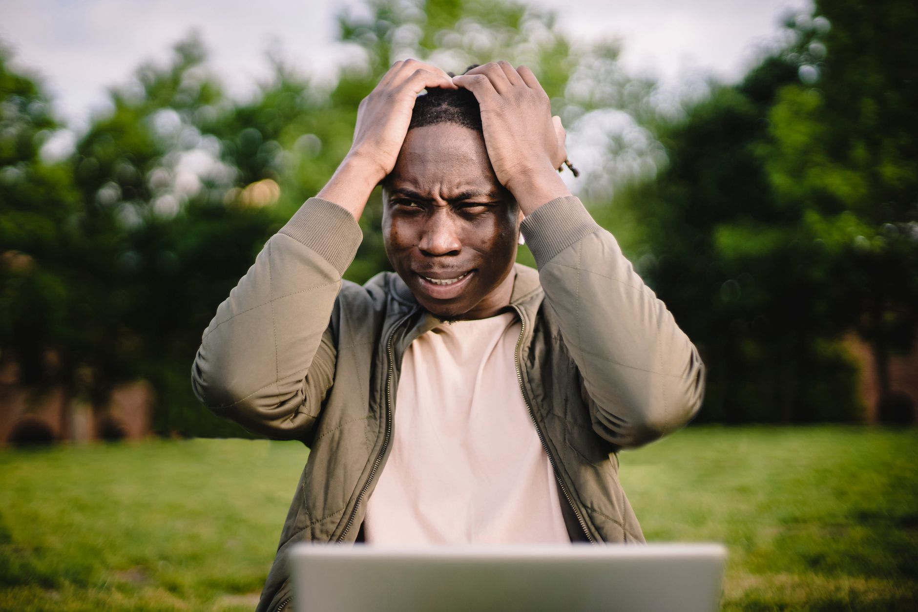 Saddened ethnic male in casual outfit holding head thinking about bad news about project while working remotely alone on laptop in park