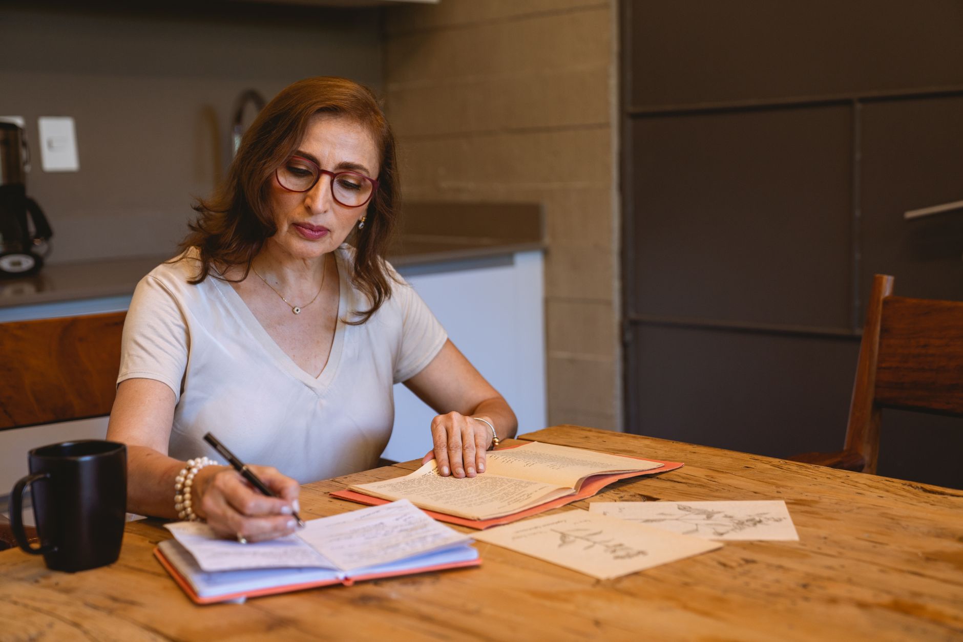 Adult woman writing notes in a cozy kitchen setting with books and a cup of coffee.